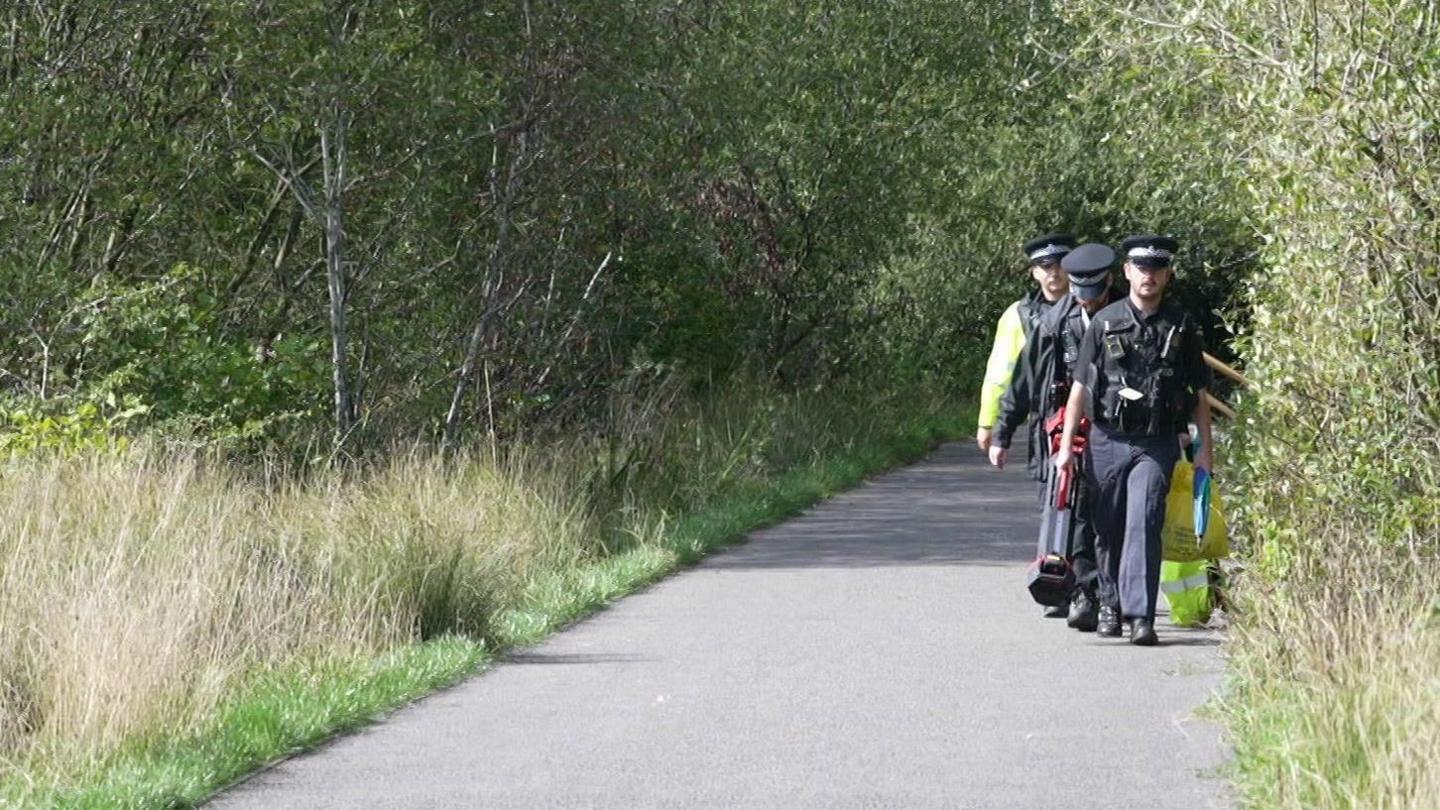 Image shows a country road with thick green foresty and vegetation either side. Three police officers are walking down the road, all wearing black uniform and hats, carrying items. One at the back has a flourescent jacket.