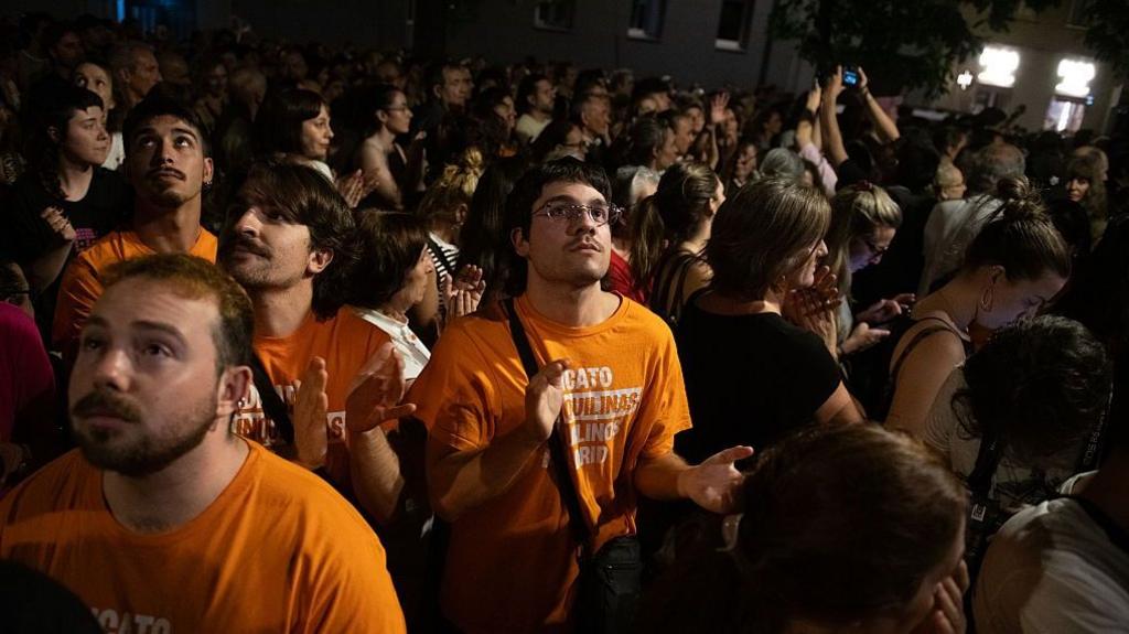 Protesters in orange tee shirts rally in a darkened street