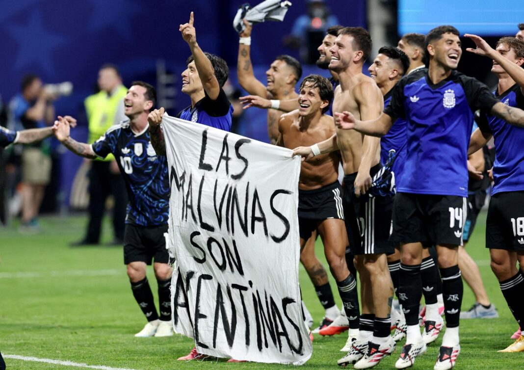 Football players, some with their shirts off, stand or jump while smiling on a green football pitch. Two of them hold a white banner that says in black writing 
