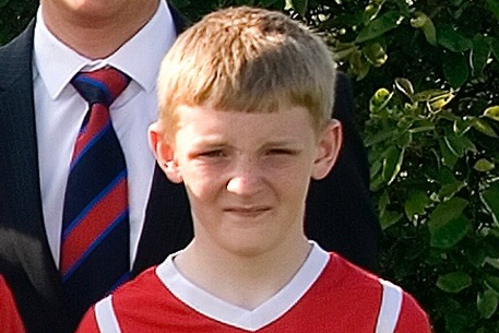 A young boy of around 14, with short blonde hair, stands looking into the camera wearing a red football kit