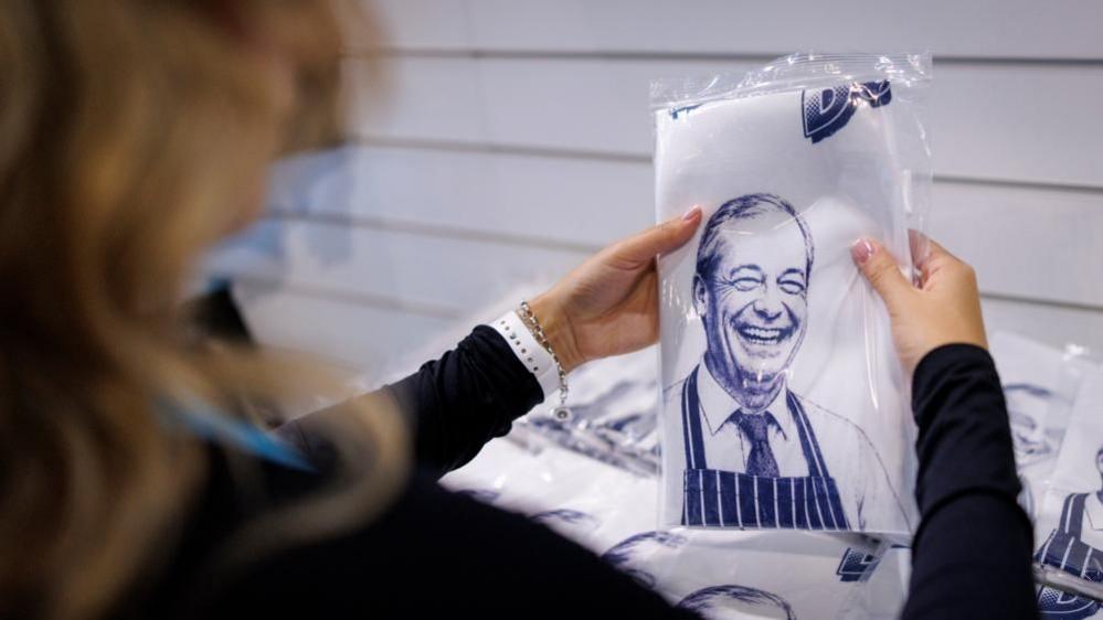 A woman with long hair and wearing a black top and a white wrist watch is holding a tea towel. The tea towel has a white backdrop and an image in blue depicting a smiling Nigel Farage, who is wearing an apron and a shirt and tie.