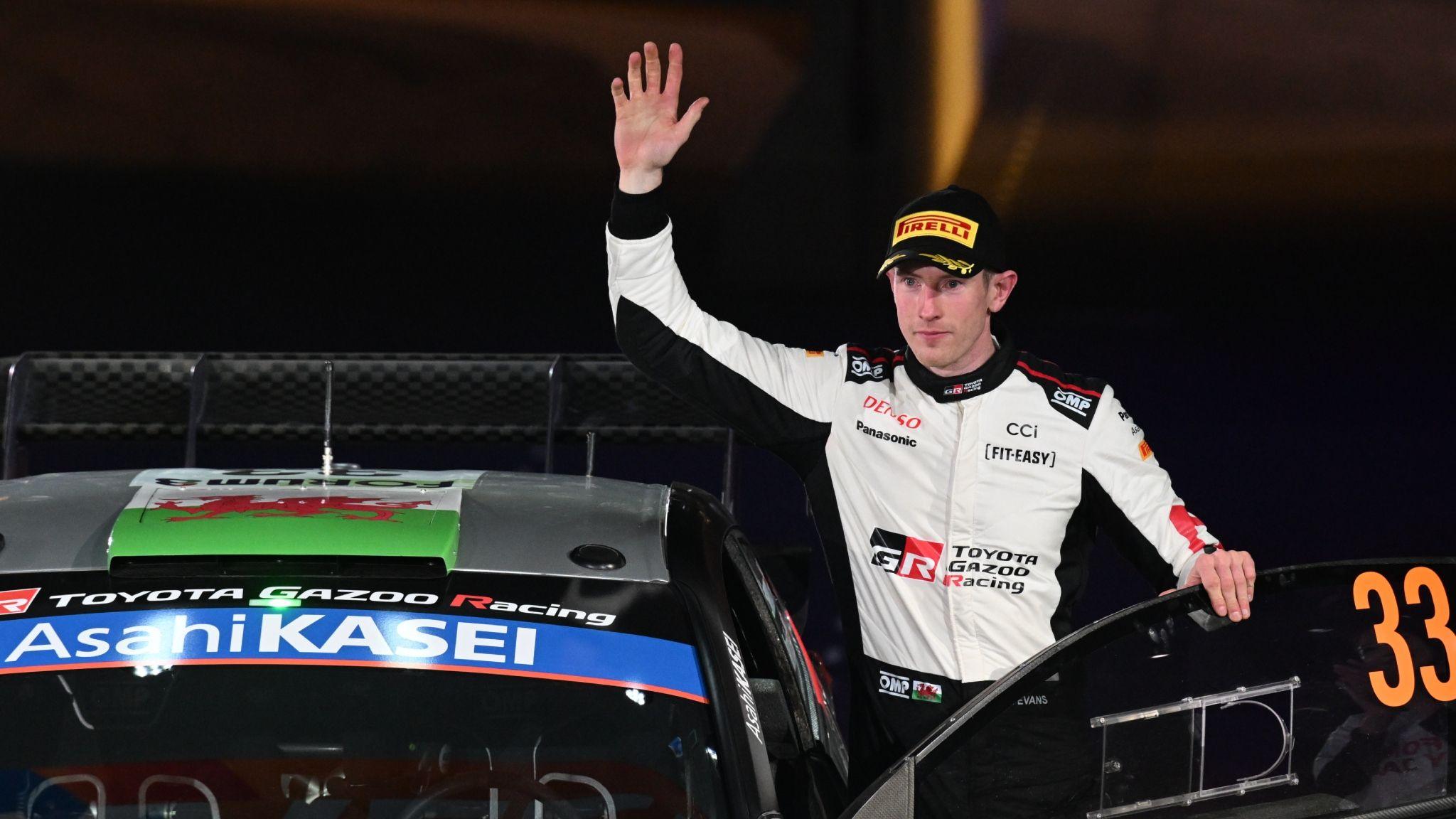 Elfyn Evans with left hand on the open door of his Toyota Yaris holds his right hand aloft