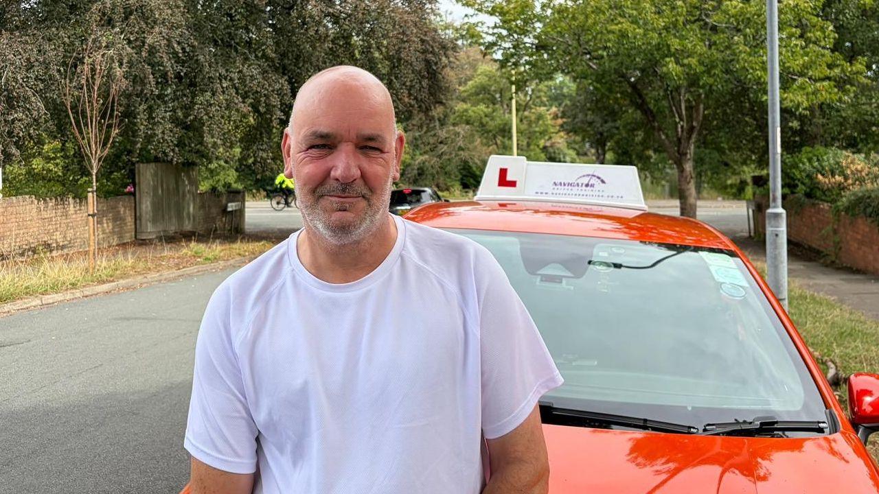 John Reynolds smiles at the camera. he is sat on the bonnet of his orange car with his driving school branding on. He is wearing a white tshirt