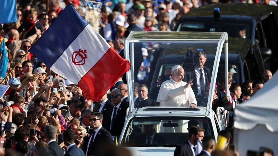 The Pope waves to crowds from the popemobile in Lourdes, as a French flag is waved