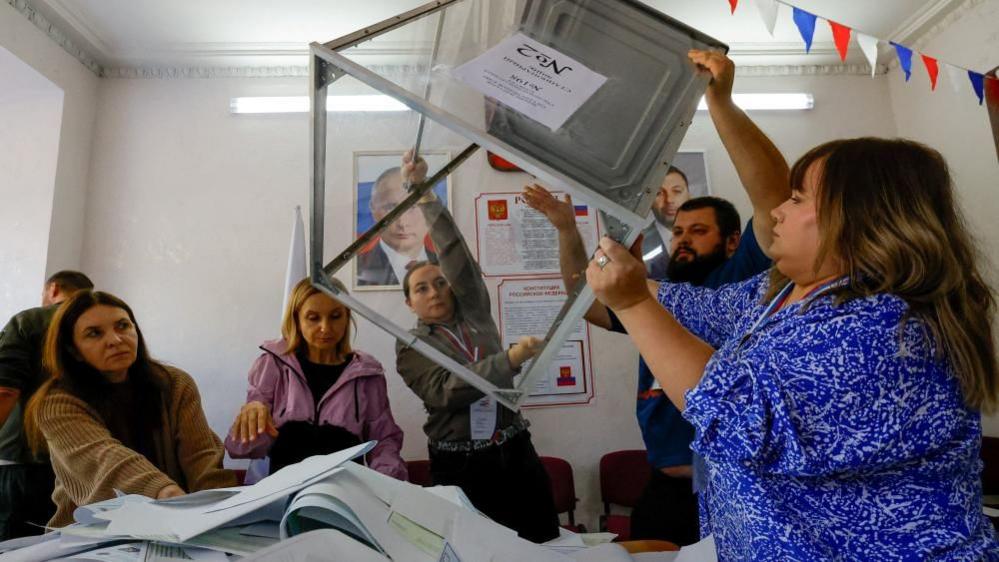 Members of a local electoral commission empty a ballot box before counting votes as they stand near portraits of Russian President Vladimir Putin