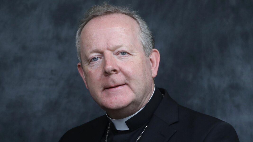 The Archbishop is looking at the camera. He has short grey hair and is wearing his religious black robes.