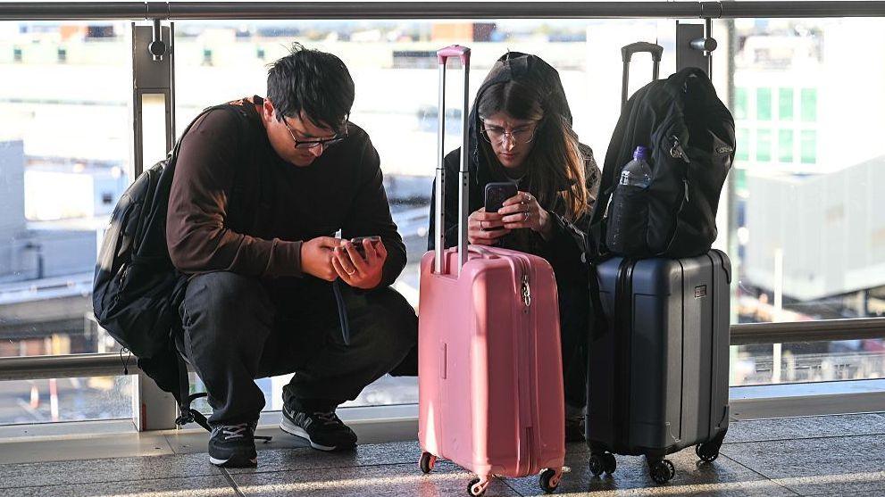 A man and a woman squat down, looking at their phones, surrounded by luggage.