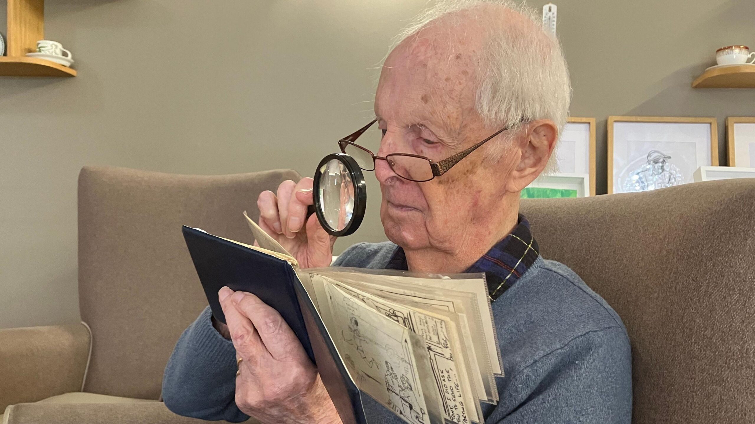 Eric Fawley is seated in a high-backed upholstered chair. He is closely examining his sketch book with the aid of a handheld magnifying glass.