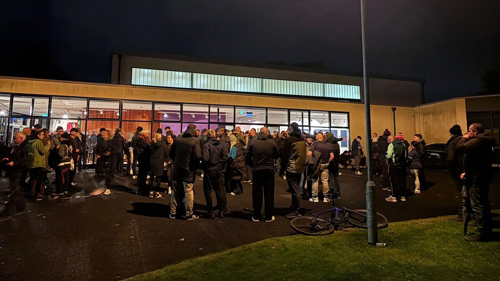 A crowd of people gather outside a church hall. It is night time.