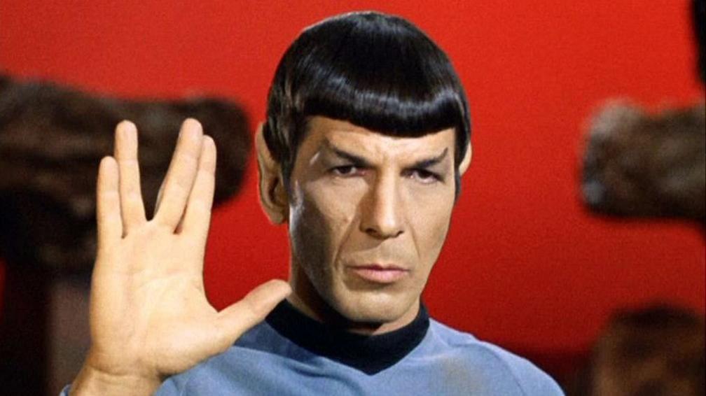 An image of one of Star Trek's main characters, Mr Spock, showing the Vulcan salute. He performs the salute by holding up his hand and parting the middle and ring finger. He is wearing a blue long sleeve with a black collar and in the background, the sky is red.