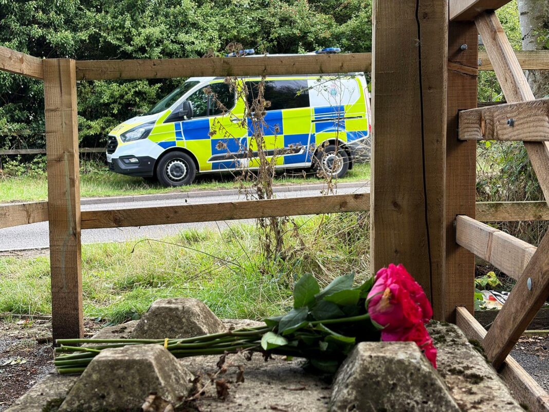 Murder arrests over baby girl’s death in woodland A bunch of roses are laid on their side by a wooden gate. In the distance is a police van parked on the grass verge next to a road.