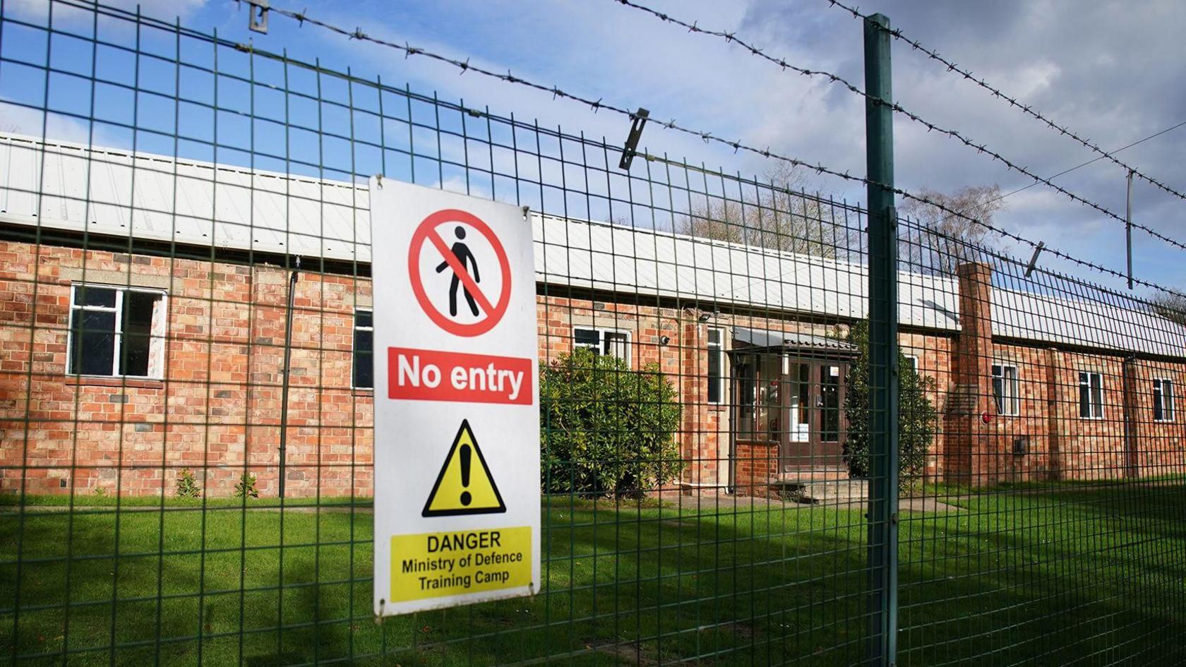 A general view of Crowborough Training Camp, East Sussex. There is a wire fence and a sign, with an area of grass behind it.