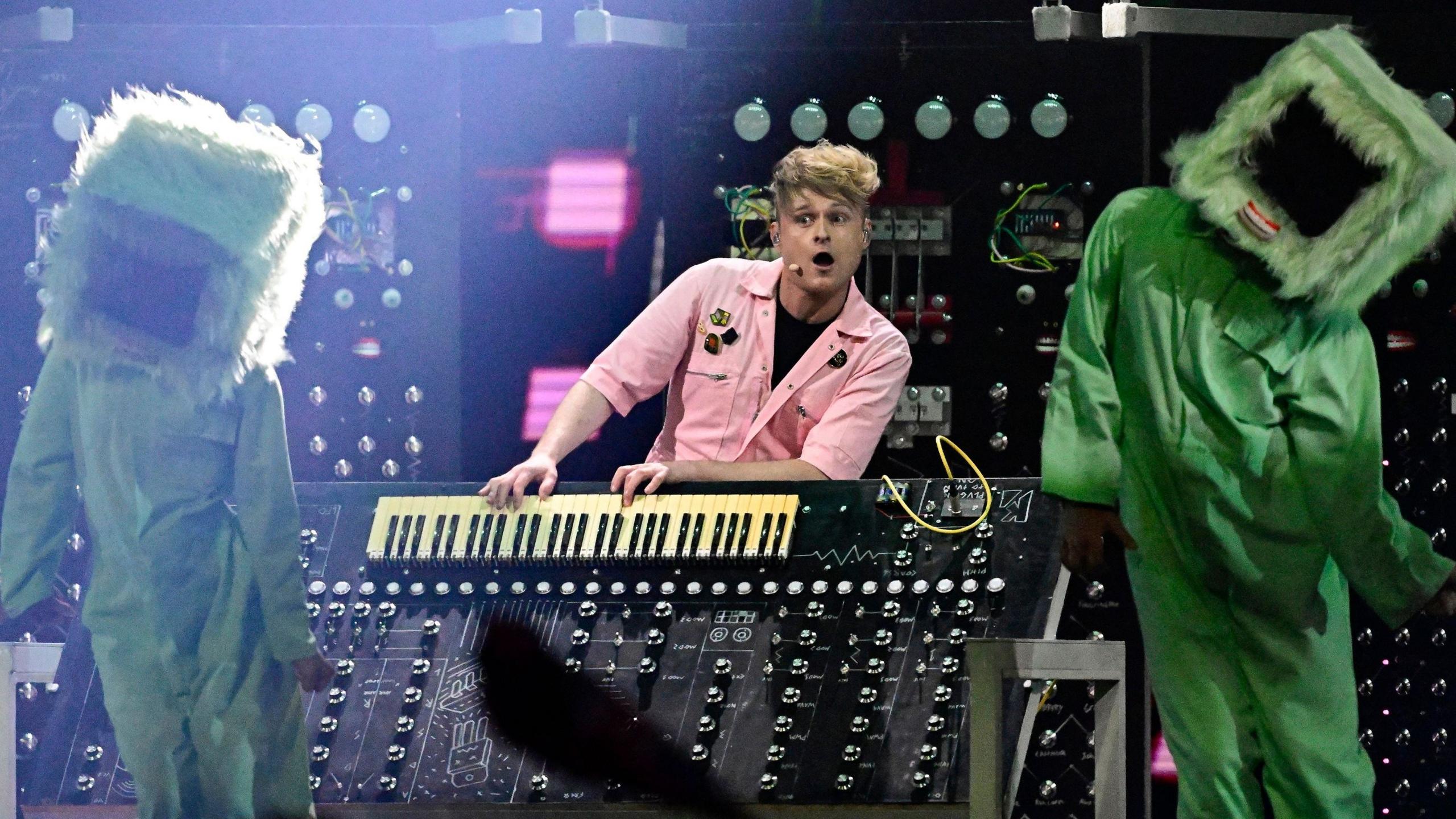 A performer in a pale pink outfit stands behind a large custom-built electronic keyboard and control console on a Eurovision stage. Two large pale green furry characters stand on either side of the instrument. Each character has a rectangular black screen in place of a face, long fluffy hair and a pale green outfit. Coloured stage lighting, electronic displays and illuminated equipment form the background while the musician plays the instrument from the centre of the stage.