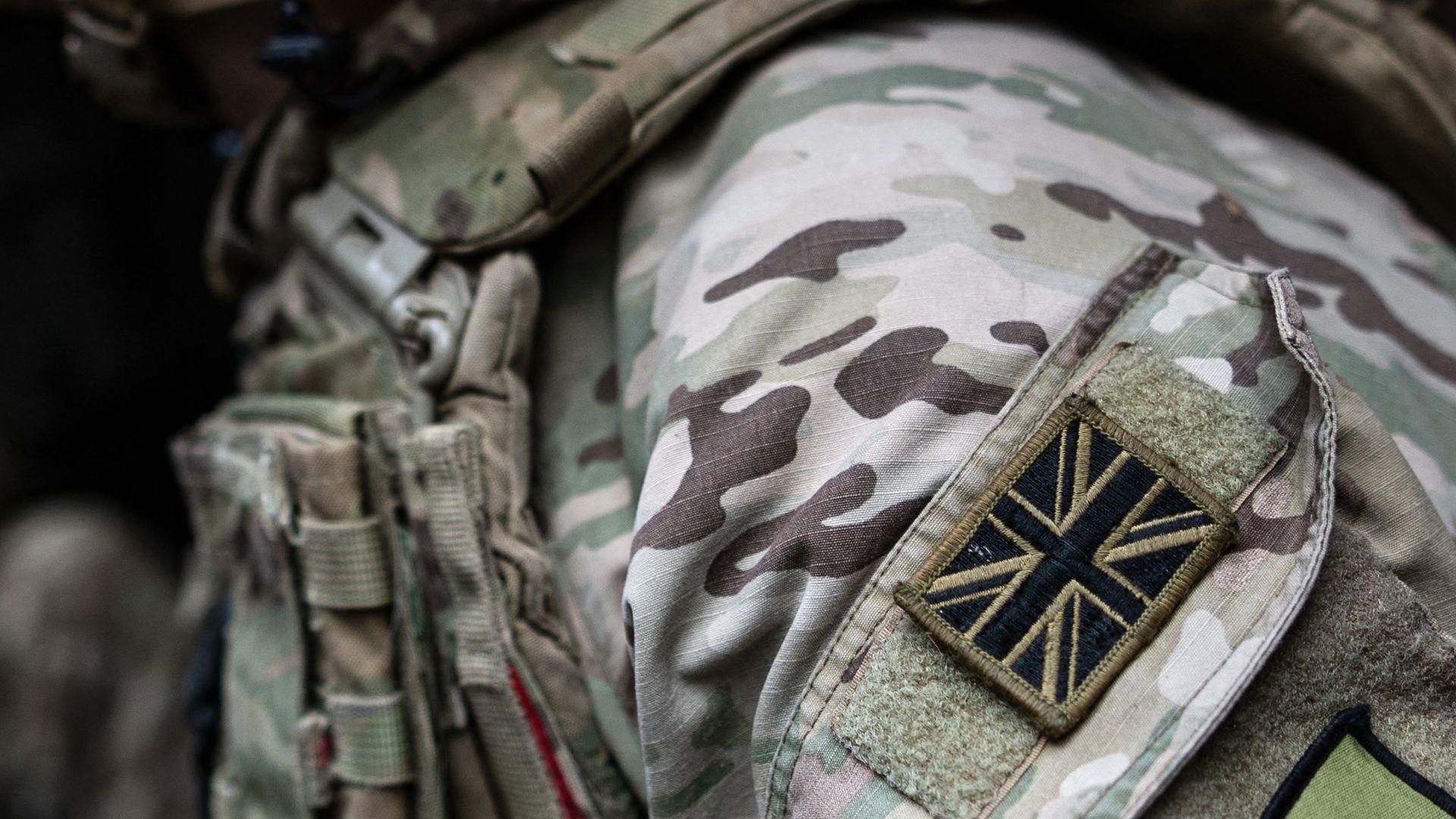 A close-up shot of a British soldier's uniform. A black and brown Union Jack patch is visible on their shoulder. The rest of the uniform is green, white and brown camouflage.