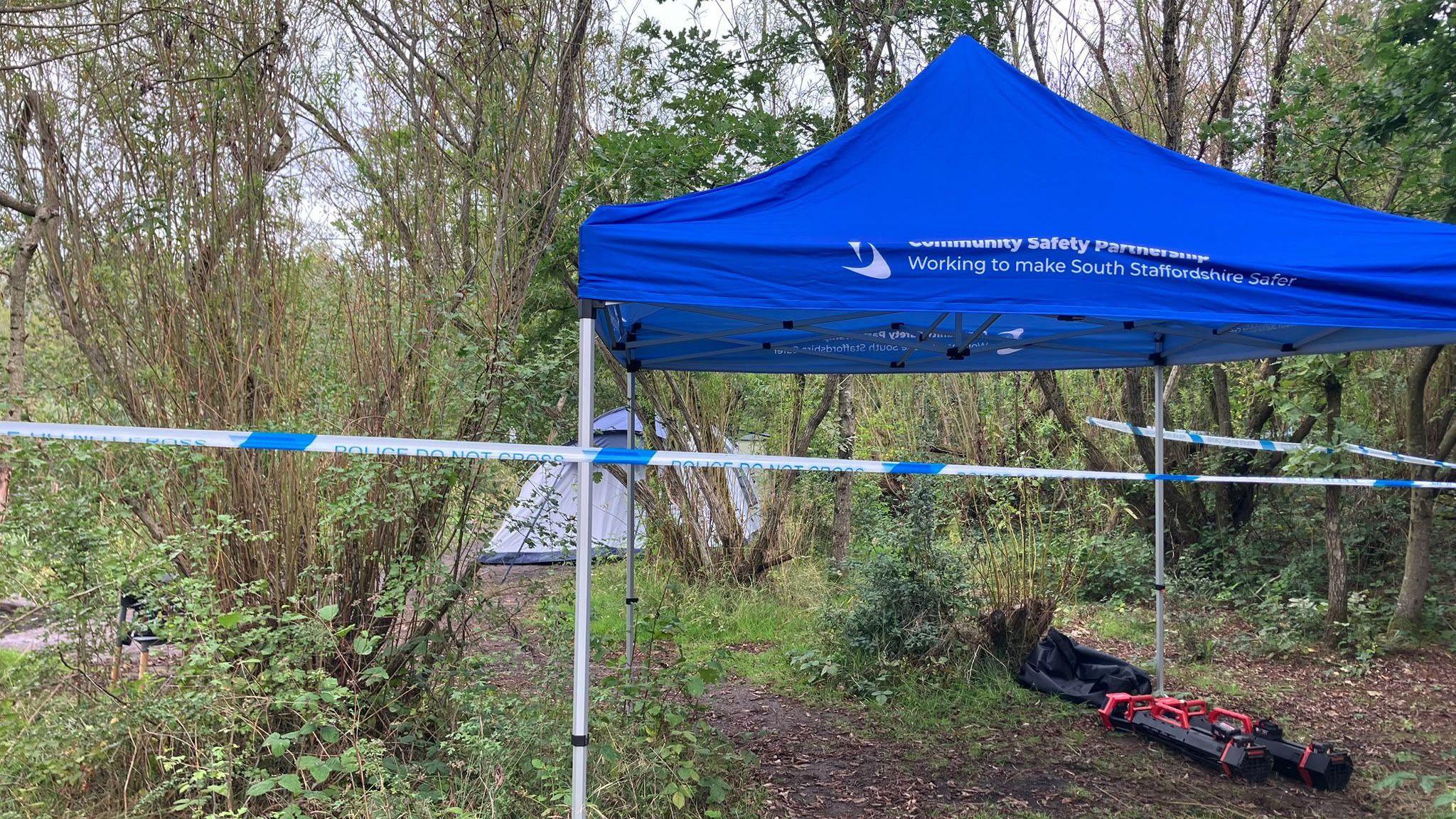 A blue gazebo with white writing on it is a woodland area. Further into the woodland is a light-coloured tent. There is blue and white police tape around the scene.