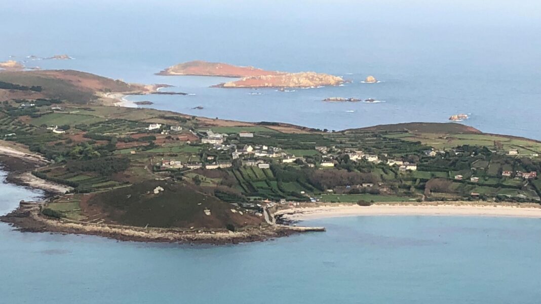 An aerial photo of St Mary's on a sunny day. The island has fields and houses and a long white sand beach can be seen. There are further small islands beyond.