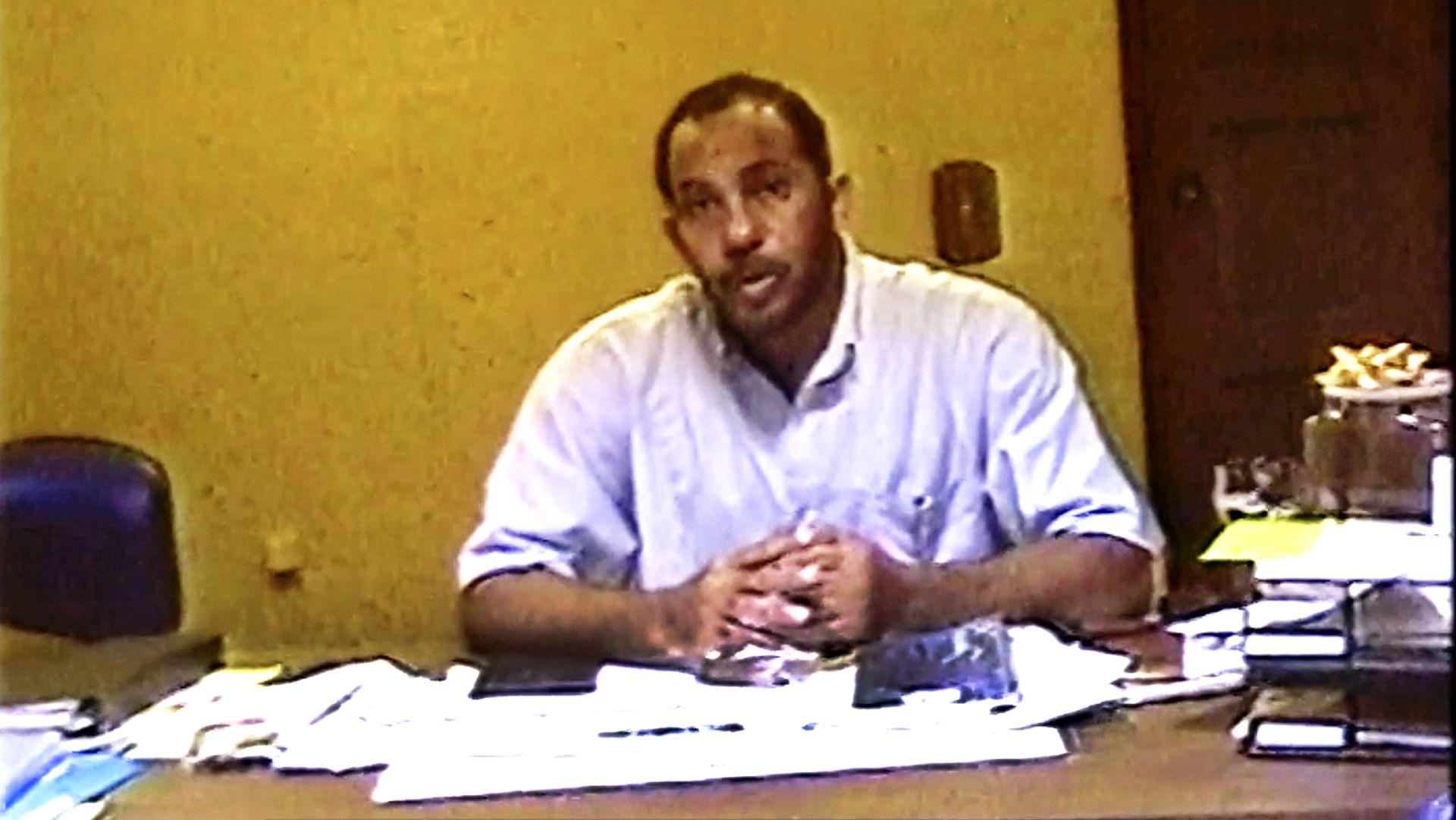 A man wearing a lilac shirt sits at a desk covered with paperwork.