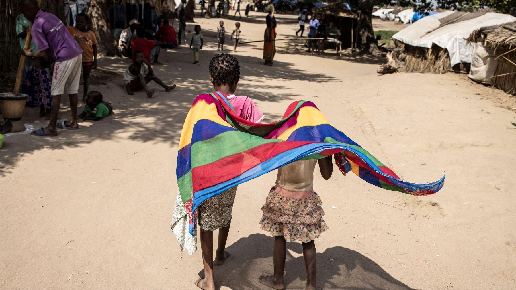 Two children with their backs to the camera with stripy coloured scarf around one of their shoulders and over the other's head as they walk down a dusty road in a displacement camp in Cabo Delgado in Mozambique.
