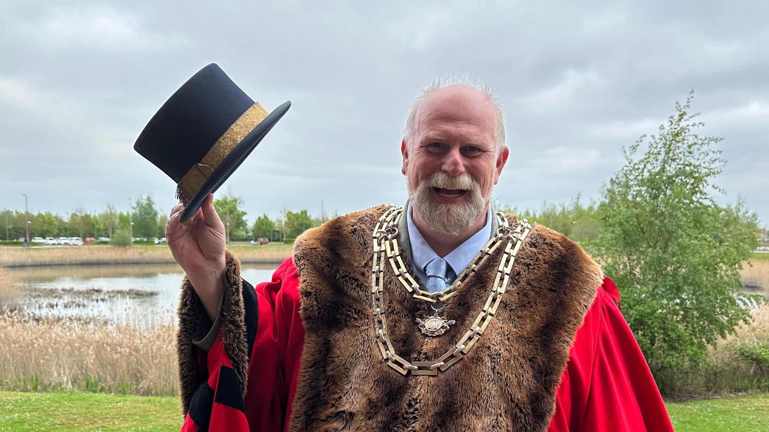 Tom Wootton wearing a bright red ceremonial robe trimmed with dark fur and a large chain of office stands outdoors beside a wetland area. He is holding a black top hat with a gold band raised in one hand. Behind him are reeds, a pond, green grass, and trees under a cloudy grey sky. The image is taken in daylight.
