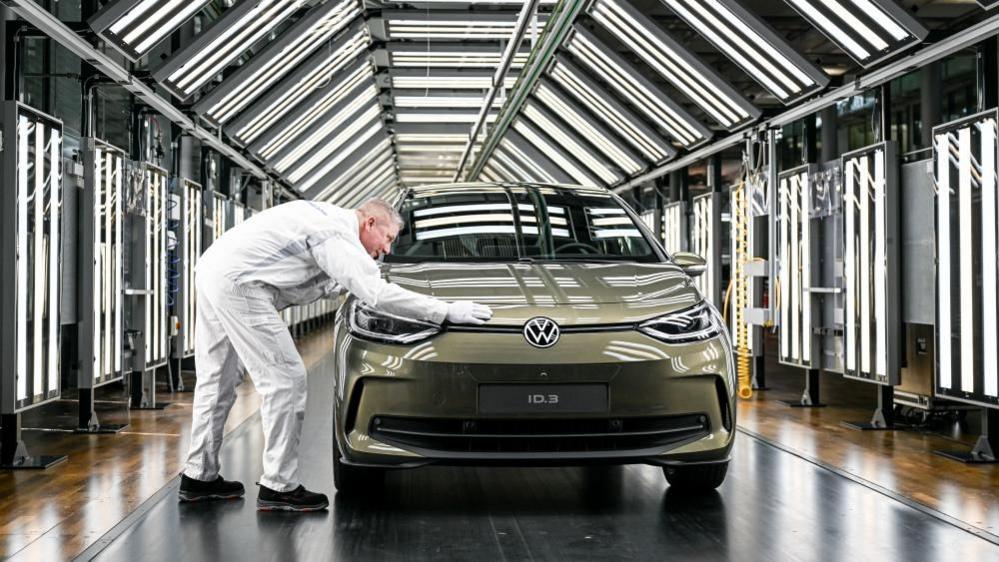 A Volkswagen employee presents a Volkswagen ID.3 car during a press presentation. He has his hand over a car, in a mossy green colour, parked in a tunnel full of white LED panels.