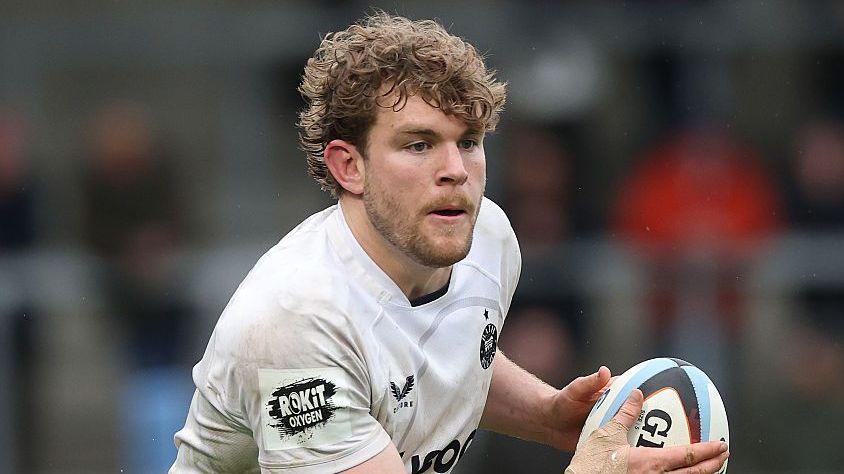 Ethan Staddon holds a rugby ball to his right in two hands as he runs on the pitch during a game last season. He is wearing a white Bath branded shirt