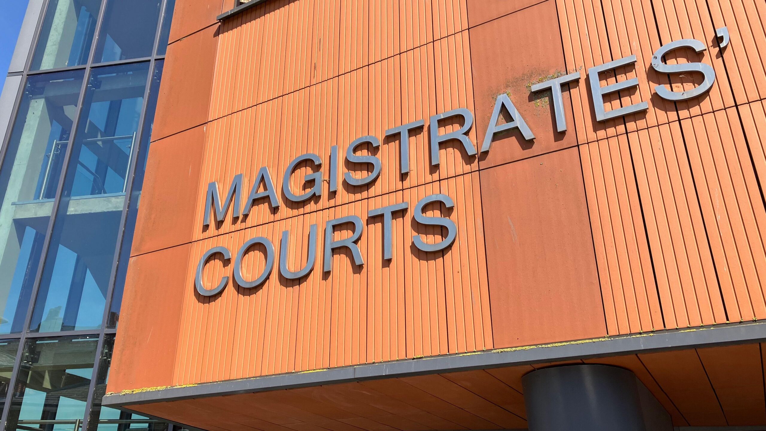 A dark orange wall at the entrance of Colchester Magistrates' Court. Large silver letters spell the words "MAGISTRATES COURTS" on the building.