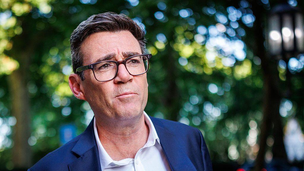 A medium close up of Andy Burnham wearing a blue suit and an open-collar white shirt with trees behind him in soft focus