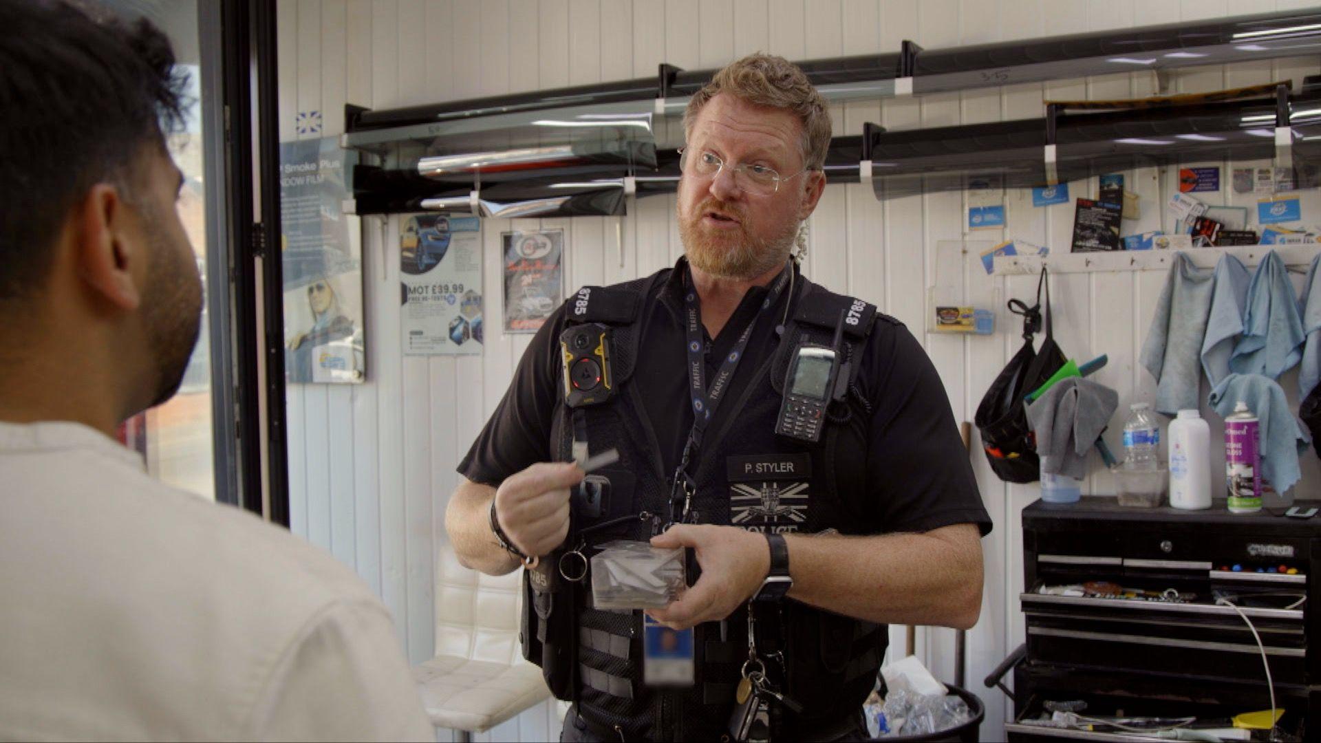 A police officer wearing a body-worn camera and radio speaks to Rahil inside a vehicle registration plate business. The officer is holding a small item while standing beside display racks and workshop equipment, with examples of vehicle registration plates mounted on the walls in the background.
