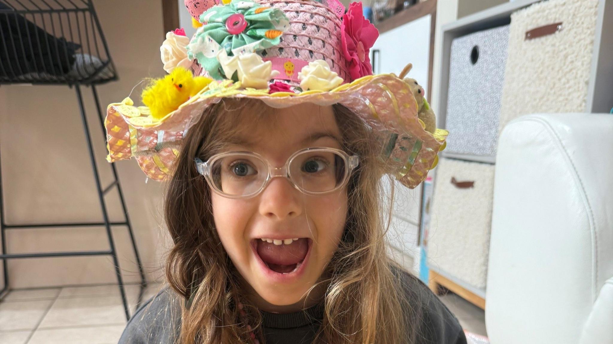 A young girl, with her mouth open, looking excited, wearing clear glasses, and an Easter hat on her head, with chicks, flowers and bright colours on her head. She is by boxes in a room with a chair to the left.