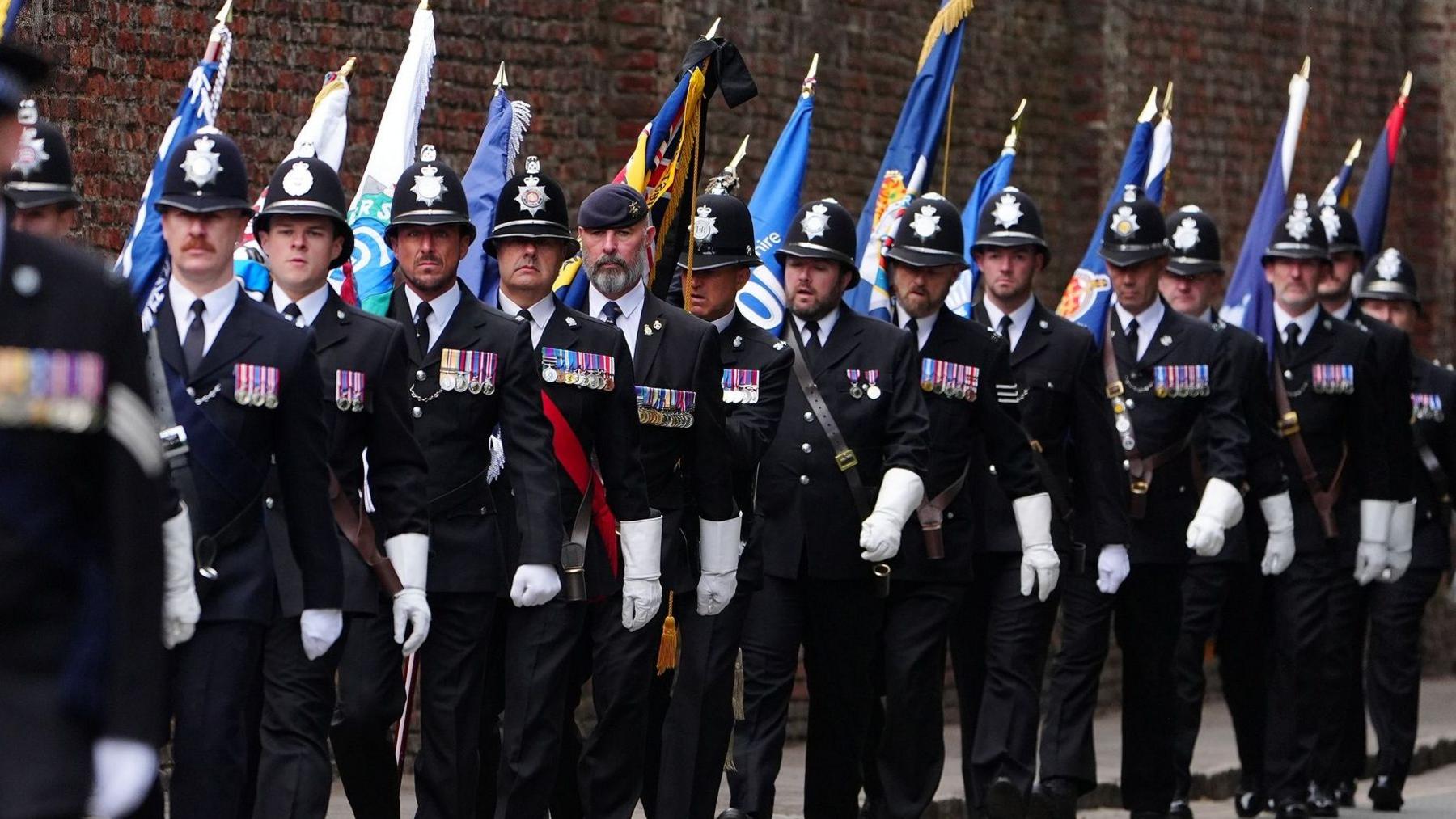 Several police officers in black uniforms with white gloves in a line while walking towards the camera. They are all men and most are wearing custodian helmets, but one man is wearing a blue army beret. Each officer has a number of colourful medals on the right breast of their blazers. There is another officer closer to the camera on the left who is out of focus. Many of them are carrying colourful flags. There is a large brick wall behind them.