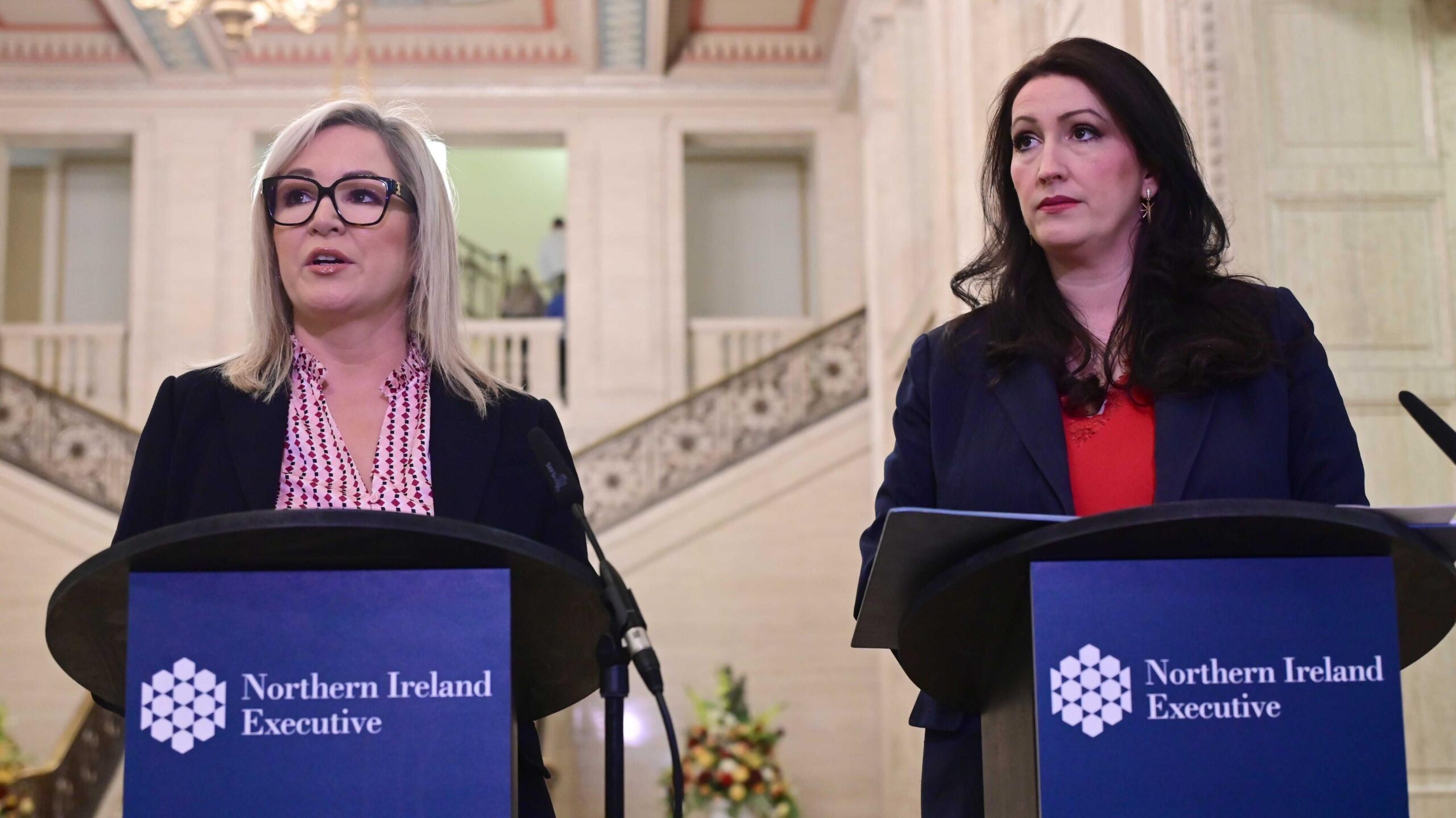 Michelle O'Neill and Emma Little-Pengelly are standing behind podiums that say Northern Ireland Exectuvie. They're in the hall at Stormont. A marble staircase is behind them.