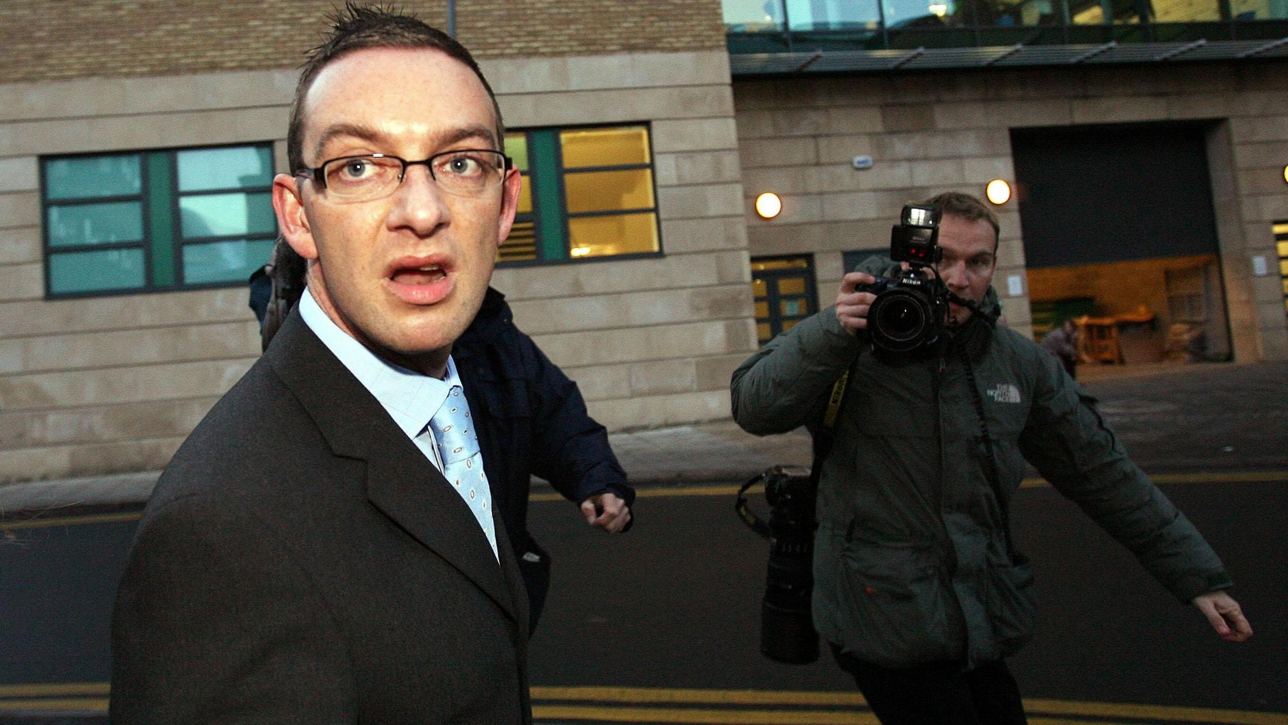 Colin Norris outside Newcastle Crown Court in 2008 - A person in a suit and glasses walks outside a building while a photographer points a camera with flash attachment towards them on a city street.