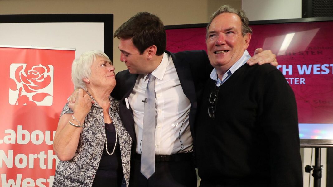 Andy Burnham hugs his parents, Eileen and Roy, after he was selected as the Labour candidate to become the mayor of Greater Manchester in September 2026. They are standing in front of a red banner reading 