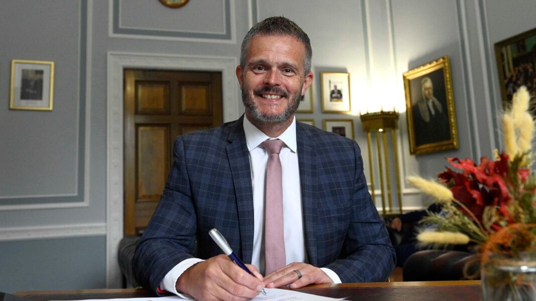 What big issues are on the agenda as Stormont returns after summer? Robbie Butler is sitting at a desk and holding a pen over a sheet of paper. He's smiling and wearing a navy checked suit with a white shirt and a soft pink tie. He's in a room with photos and portraits hanging on the walls.