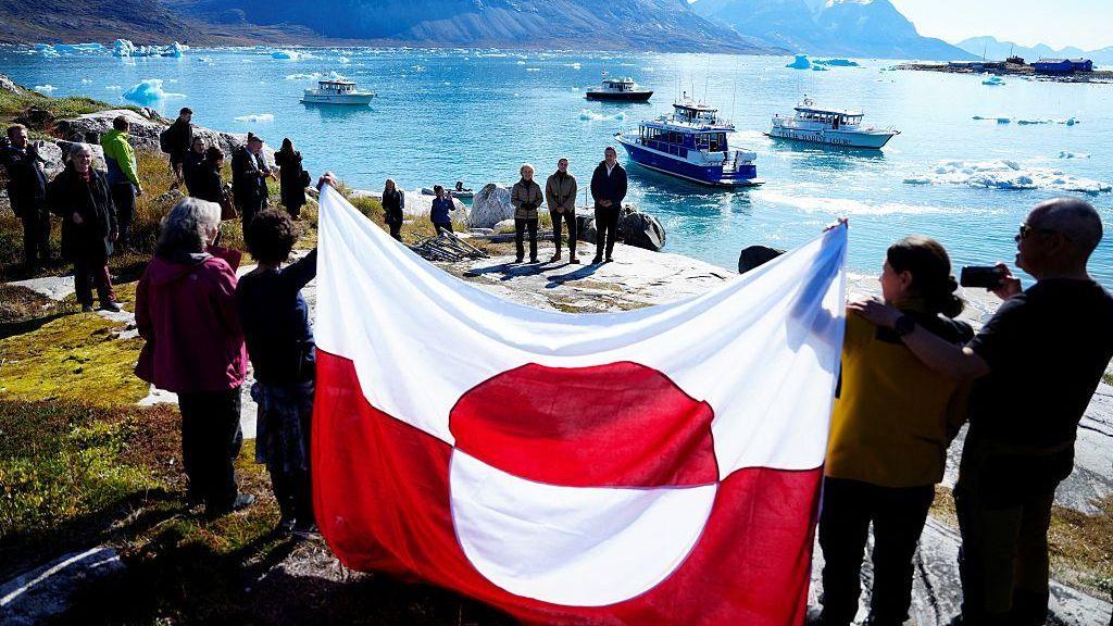 People hold a Greenland flag on the shoreline of Bear Island with boats floating in the background on a sea spotted with large chunks of ice