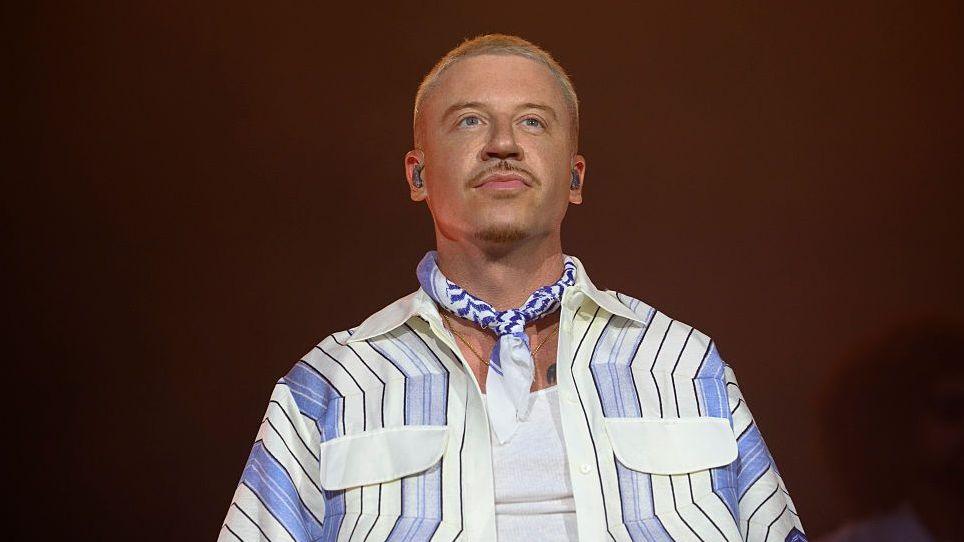 Macklemore on stage, he has cropped blonde hair and is wearing a white vest and pale blue, white and black striped shirt. Around his neck he is wearing a blue and white neckerchief.