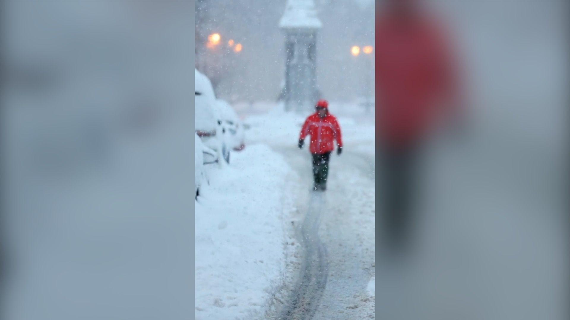 A man in the distance walks up a road covered in snow in a red coat with his hood up.