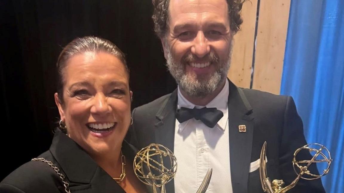 Rachel wears a black suit and gold necklace, her hair tied back. Matthew wears a black suit and white shirt. They are smiling and holding two Emmy awards