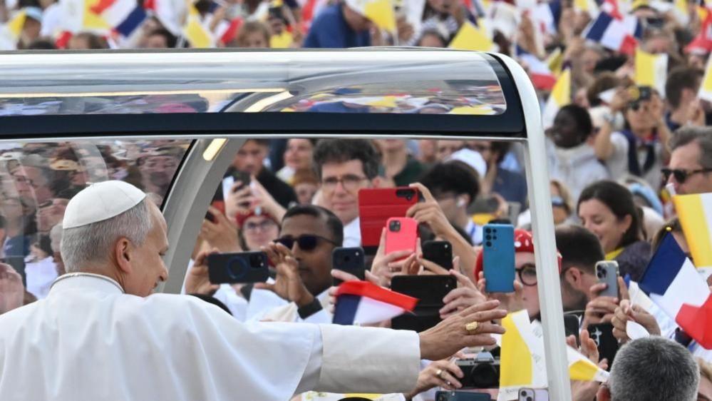 The pope waves to the crowd from his popemobile. A group of people all holding their phones up can be seen in front of him