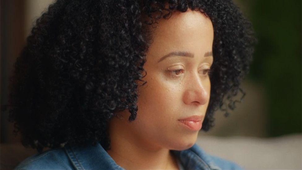Close-up portrait of Annie, who has dark curly hair and is wearing a blue denim shirt, photographed indoors. Annie is shown in profile, looking down and away from the camera. The background is softly blurred, with warm lighting and muted green and beige tones visible behind her.