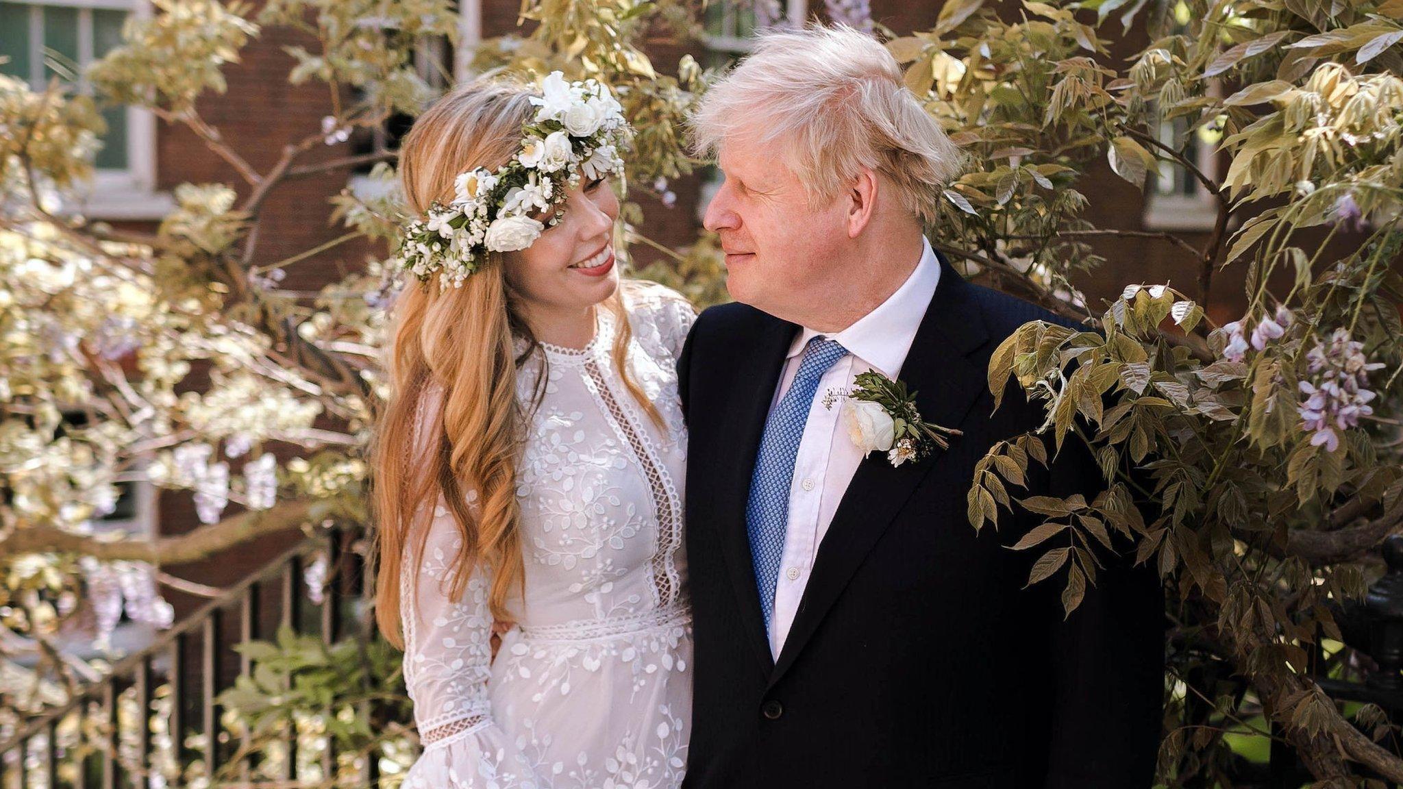 Boris Johnson standing next to his wife Carrie with his arm around her. He is wearing a black suit, white shirt and blue tie. Carrie is wearing a lace wedding dress with intricate detailing down the middle and on the sleeves. She is also wearing a white rose flower crown and smiling at her husband. They are standing on an outdoor staircase surrounded by foliage and flowers.