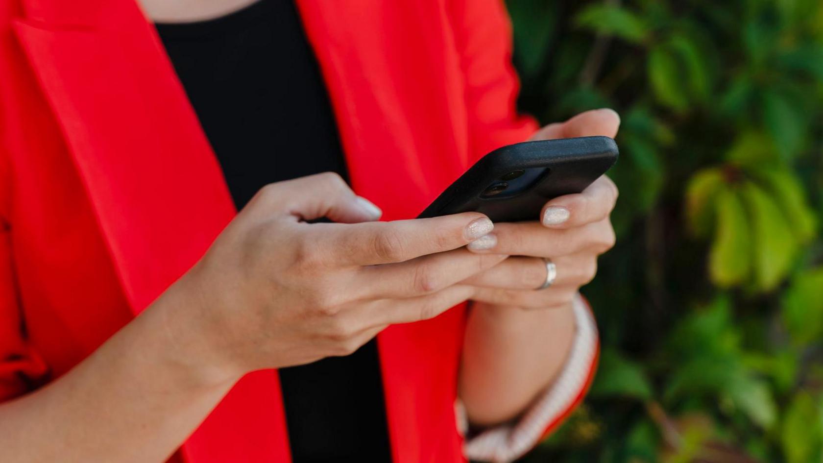 Close up of a woman's hands holding a black smartphone. She is wearing a bright red jacket and a silver ring.