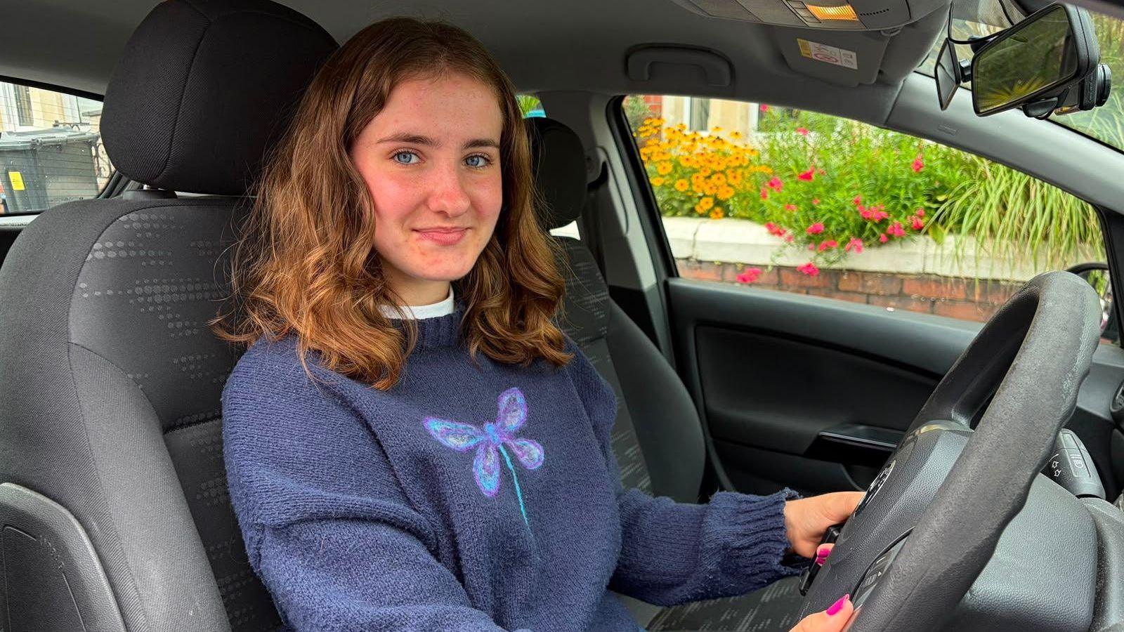 Megan sits in her car. she is smiling at the camera and has reddish to brown hair cut at her shoulders. She is wearing a blue jumper with a dragonfly on.