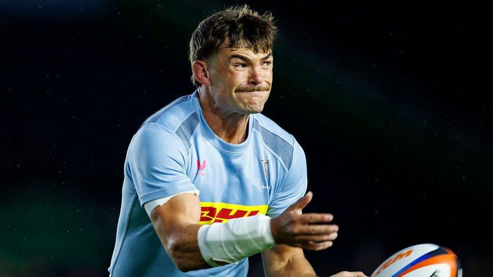 George Furbank holds his hands out after throwing a rugby ball to his left. He is wearing a light blue Harlequins shirt