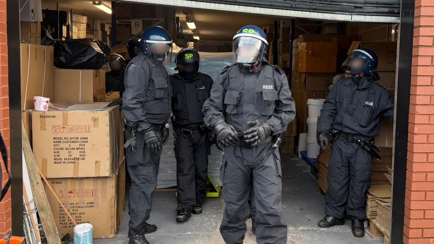 A group of four police officers in all-black uniforms and with helmets and masks are standing in a warehouse section of a red brick building.