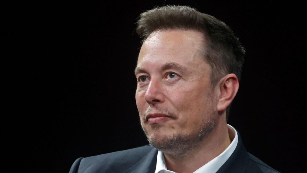 A headshot of Elon Musk looking off camera, wearing a black suit jacket and white shirt. He has stubble and brown hair
