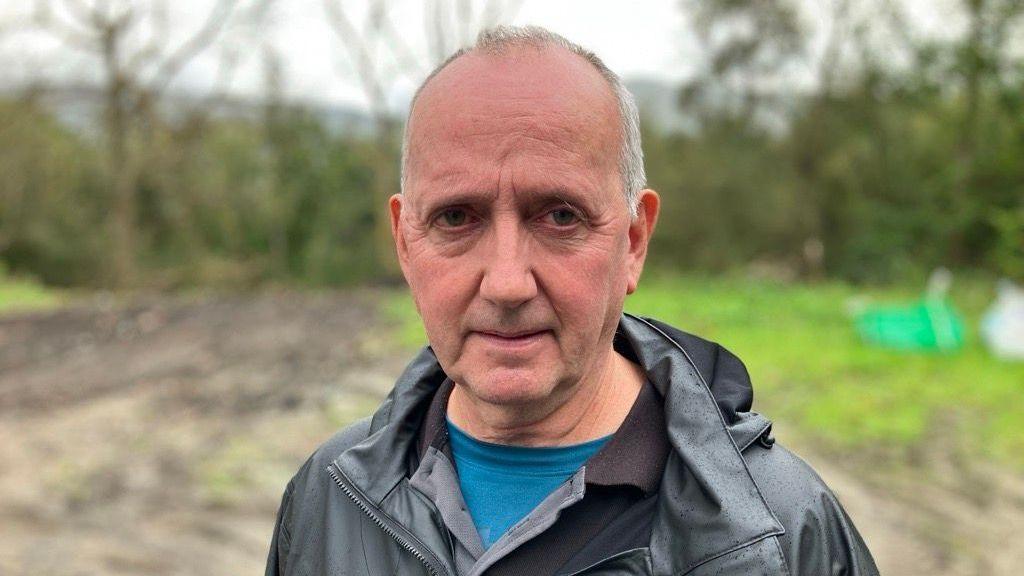 A white man with short grey hair. He is wearing a black leather coat, and a blue t-shirt underneath. In the background is a field with patches of grass.