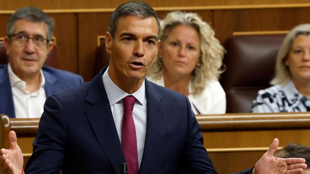 Spanish Prime Minister Pedro Sánchez addresses lawmakers in parliament, speaking at a lectern with hands outstretched, while several seated politicians look on behind him