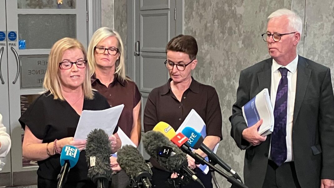 Diane has shoulder length blonde hair and is wearing a black dress. She is speaking into a mic and has rectangular glasses. There are three other people standing beside her who are in shot.