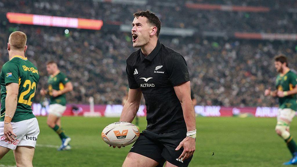Will Jordan, in New Zealand's all-black kit, celebrates with a satisfied facial expression and with the ball in his right hand after scoring against South Africa